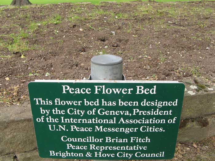 Picture of a raised flower bed in a park. The sign reads 'Peace flower bed.' The bed behind is empty exept for weeds.