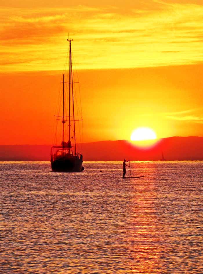 A man standing on a surfboard punts across the sea with an orange sunset behind him and a sailboat anchored behind him.