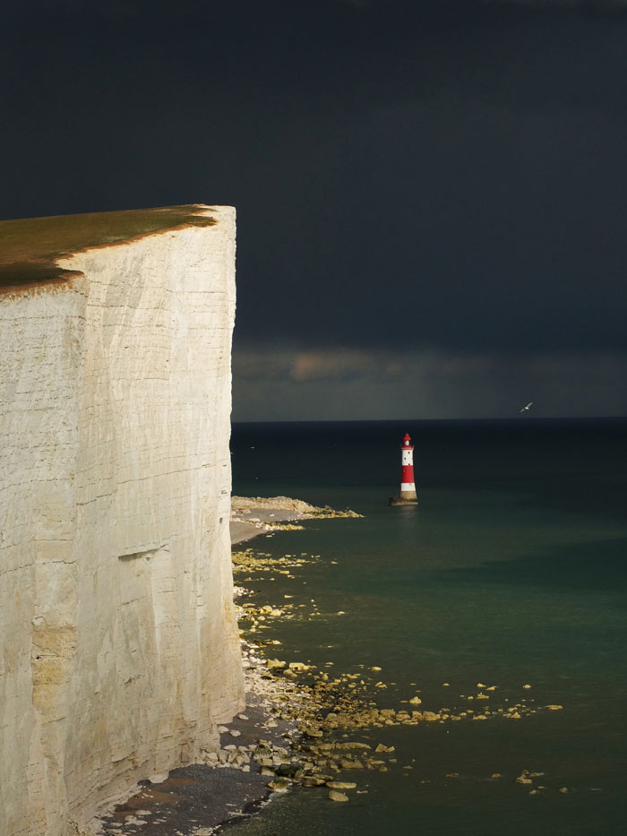 A lighthouse with a dark storm approaching and white cliffs in the foreground.